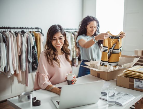Female working on a laptop while another woman unpacks clothes inside a small clothing boutique.