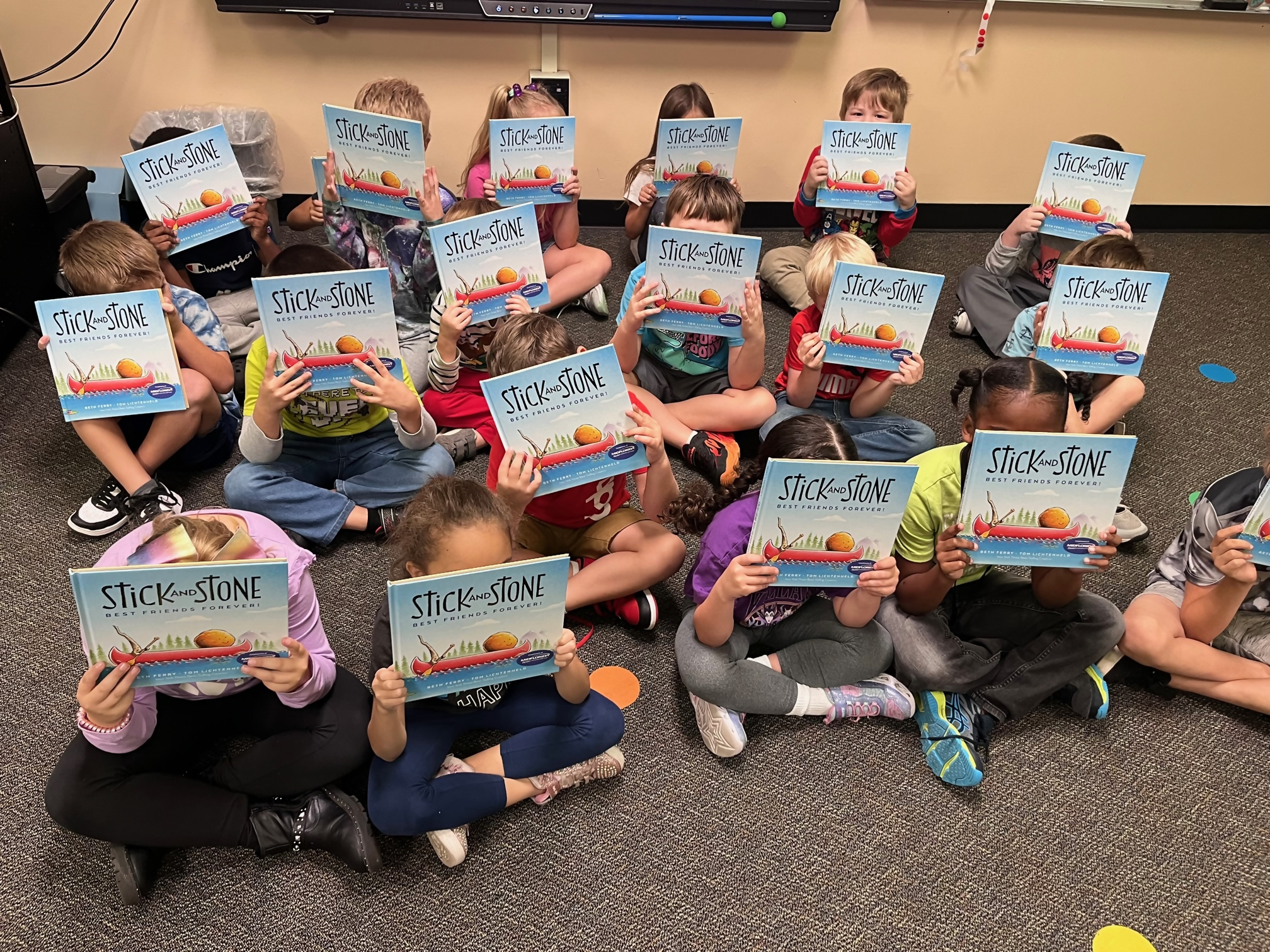 Children in classroom holiding book in front of face.