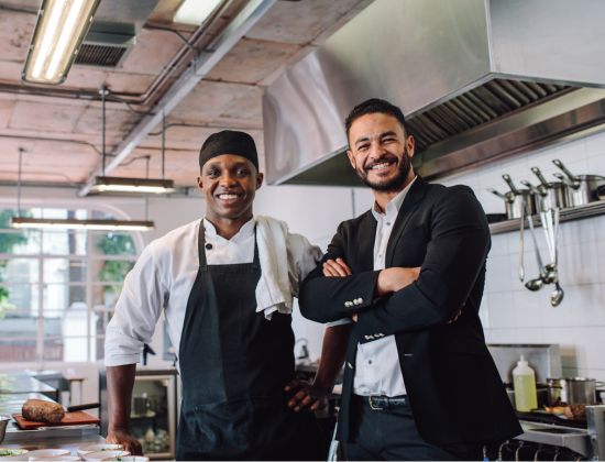Male business owner in suite standing with chef in a commercial kitchen, both smiling.