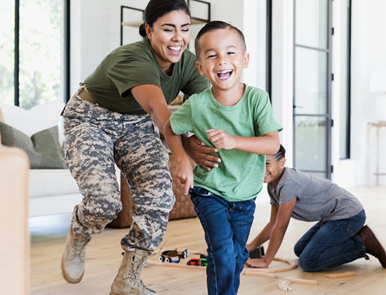 While the preteen boy sets up the wooden train set on the living room floor, the female soldier chases the elementary age boy through the house. Everybody is smiling and happy.