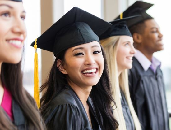 Group of four college graduates standing in a line with young woman smiling at camera.