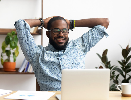 Smiling Afro-American businessman holding hands behind head sitting at office desk behind laptop.