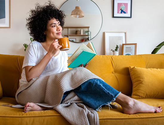 Relaxed African American woman reading a book at home, drinking coffee sitting on the couch. 