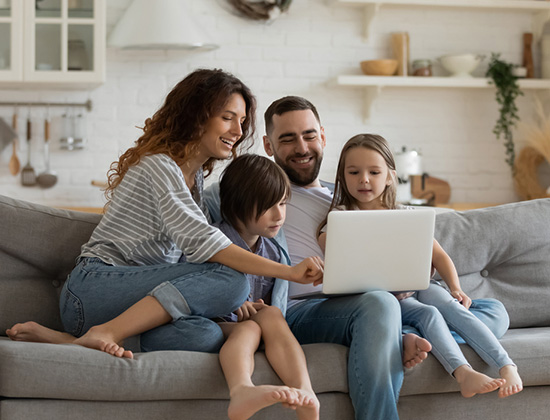 Happy young family with little kids sit on sofa in kitchen have fun using modern laptop together, smiling parents rest on couch enjoy weekend with small children laugh watch video on computer at home