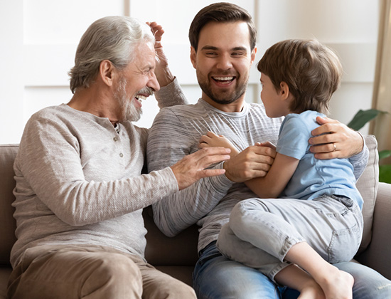 Happy three generations of men sit relax on couch in living room having fun together, overjoyed small preschooler boy play with young dad and elderly grandfather, enjoy family weekend at home