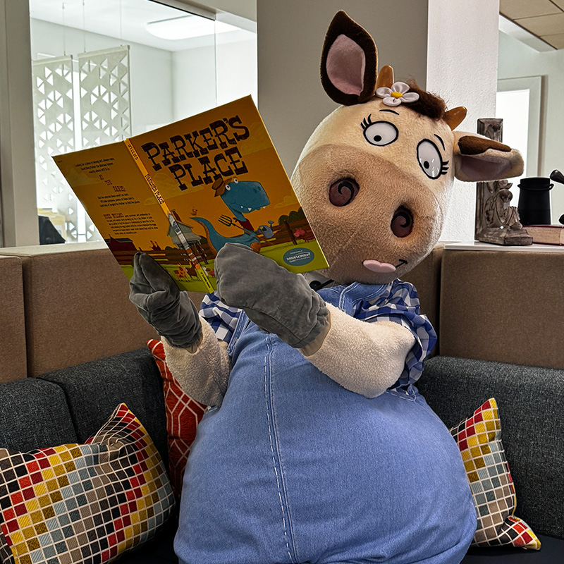 Penny the cow mascot sitting on couch looking at camera while reading the children's book Parker's Place