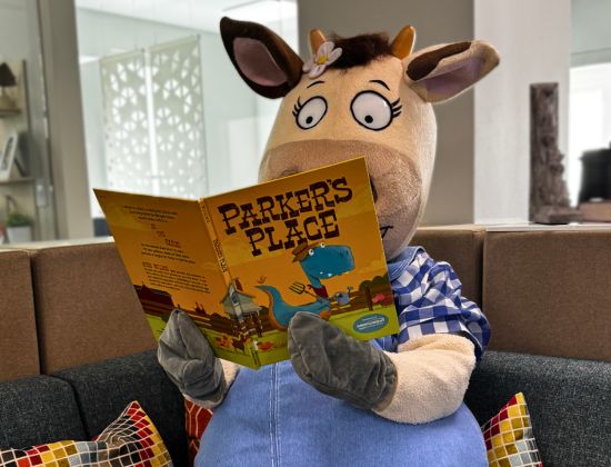 Penny the cow mascot sitting on couch reading the children's book Parker's Place