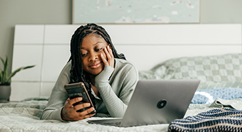 girl laying on bed looking at laptop and phone