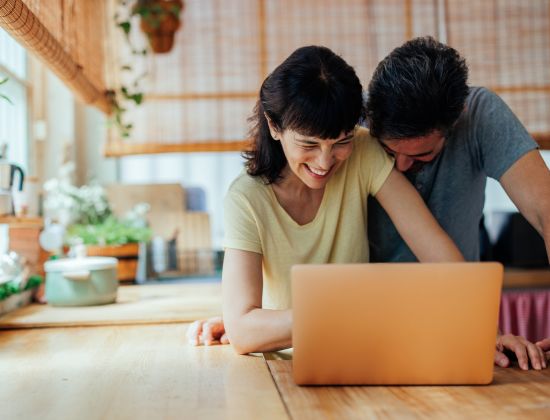 Couple on laptop in kitchen laughing.