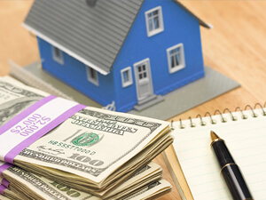 A stack of money sitting next to a miniature house with a pen and notepad