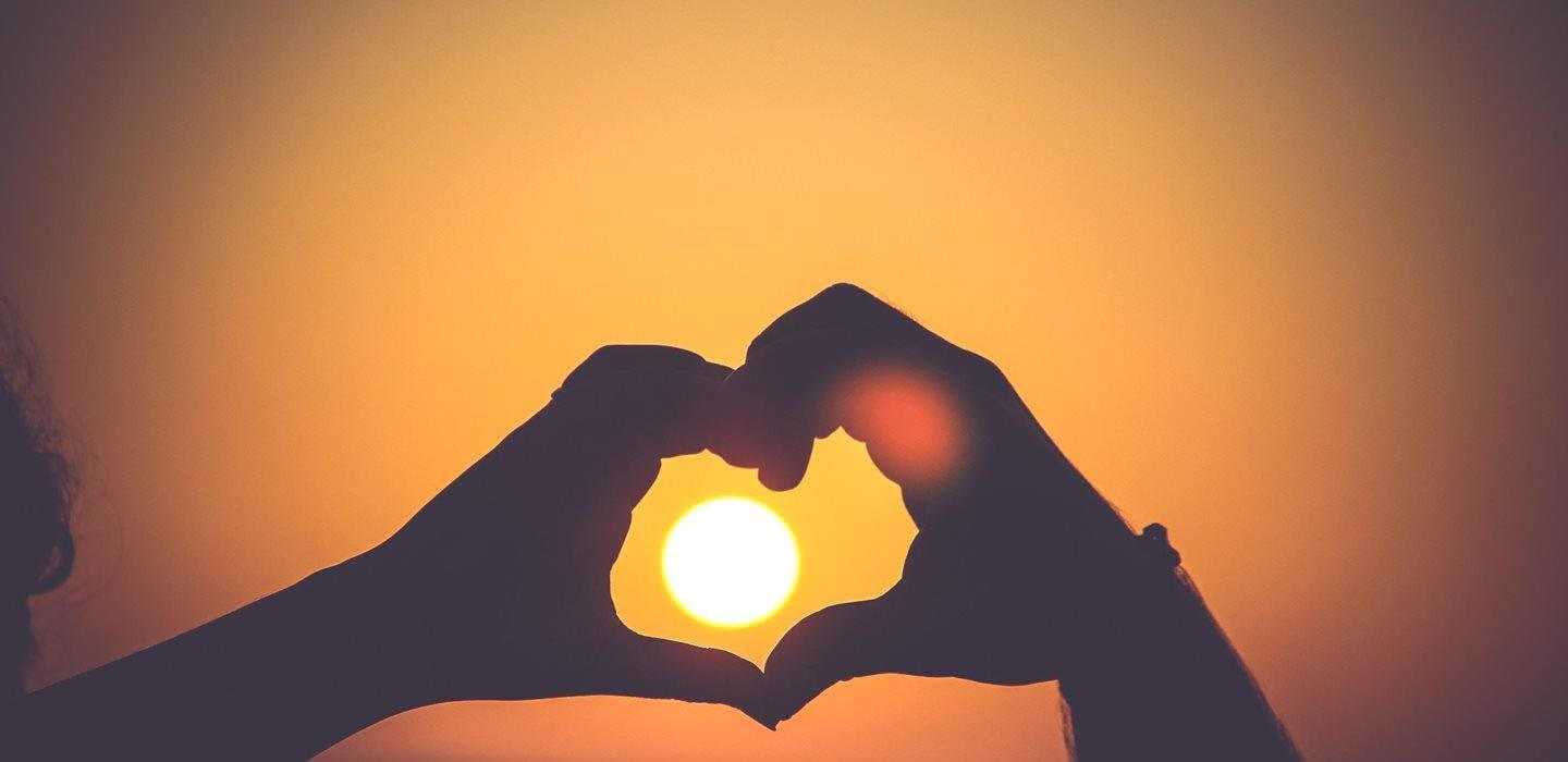 Two people standing by the sun with their hands together heart shaped over the sunshine
