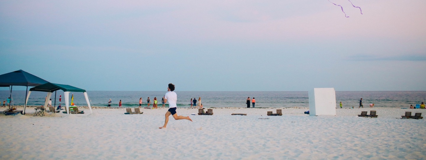 A man running while flying a kite on a crowded beach surrounded by water and blue skies